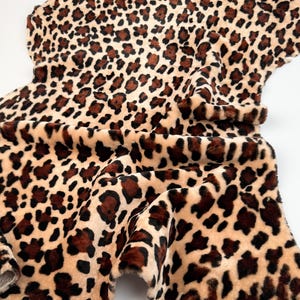 May include: A faux fur fabric with a leopard print pattern. The fabric features a tan background with dark brown spots. The material is soft and has a textured appearance, with folds and curves visible throughout the image.