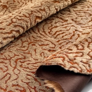 May include: Close-up of a textured fabric with a swirling pattern in shades of tan and brown. The fabric has a soft, fur-like appearance with a dark brown leather backing visible in the folds. The overall effect is of a luxurious material.