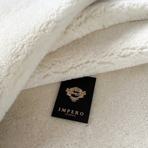 May include: A close-up of a white faux fur throw blanket with a black label that reads "Impero London".