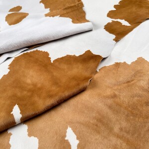 May include: Close-up of a cowhide rug with a natural pattern. The rug features a white background with large, irregular brown patches. The texture of the hair is visible, and the hide is folded in places, showing the underside.