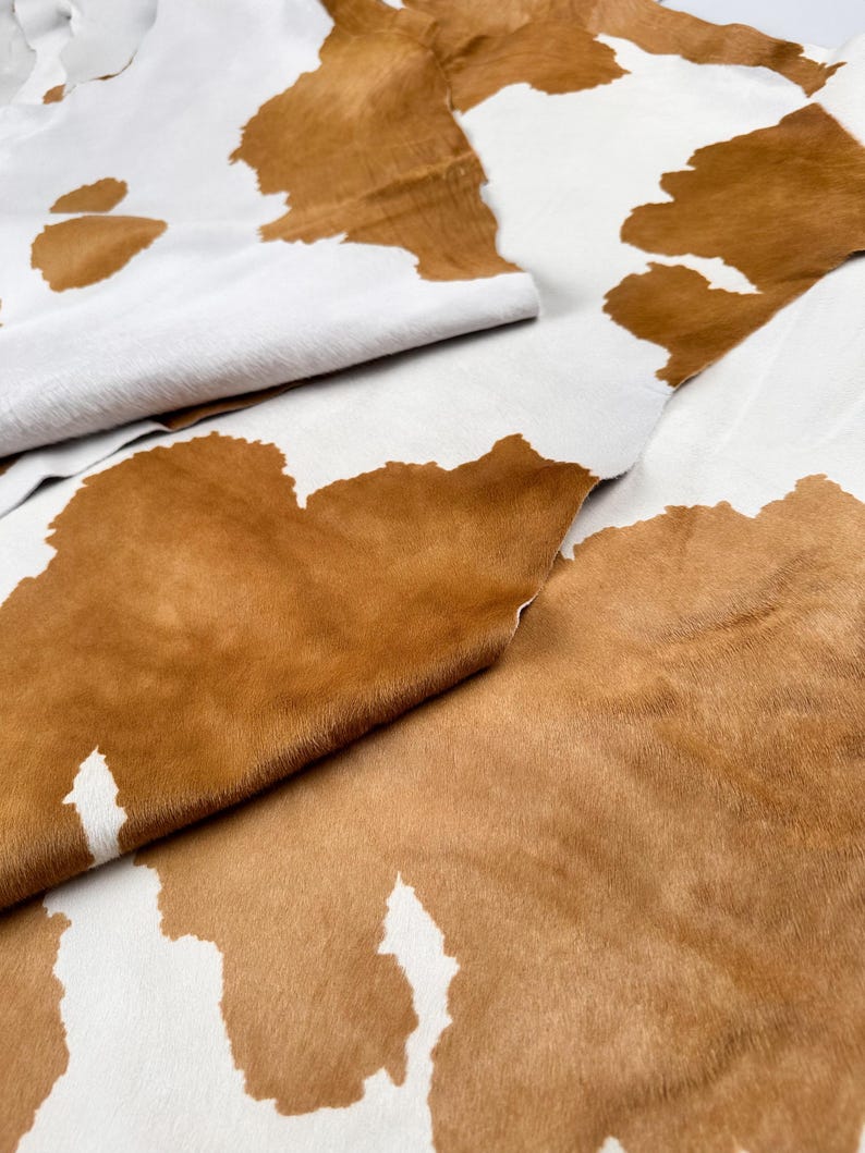 May include: Close-up of a cowhide rug with a natural pattern. The rug features a white background with large, irregular brown patches. The texture of the hair is visible, and the hide is folded in places, showing the underside.