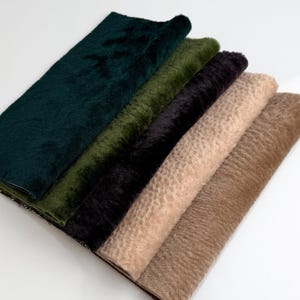 Op de afbeelding: Een selectie van imitatiebont stofstalen in verschillende kleuren, waaronder donkergroen, olijfgroen, zwart, lichtbeige en bruin. De stalen zijn diagonaal gerangschikt en tonen de textuur- en kleurvariaties.