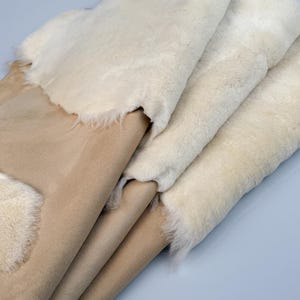 K&ouml;nnte beinhalten: Stapel aus vier Schaffellteppichen in Beige und Wei&szlig;. Die Teppiche sind gefaltet und haben eine weiche, flauschige Textur.