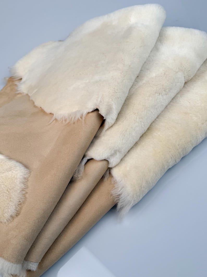 K&ouml;nnte beinhalten: Stapel aus vier Schaffellteppichen in Beige und Wei&szlig;. Die Teppiche sind gefaltet und haben eine weiche, flauschige Textur.