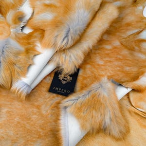 Könnte beinhalten: Eine Ansammlung von orangefarbenen und weißen Pelzfellen mit weicher Textur. Die Felle sind geschichtet und zeigen eine weiße Unterseite. Ein schwarzes Etikett mit goldenem Text trägt die Aufschrift "IMPERO LONDON".