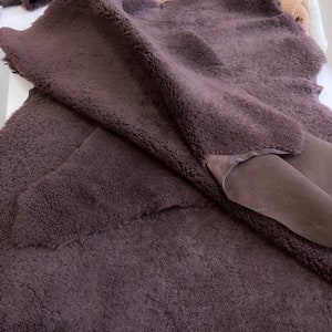 May include: Close-up of several pieces of dark brown sheepskin with a soft, textured surface. The sheepskin has a plush, fluffy appearance, with a smooth, suede-like underside. Other pieces of sheepskin in lighter shades are visible in the background.
