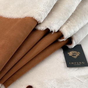 Puede incluir: Una pila de tela de cuero marrón y piel sintética blanca con una etiqueta negra que dice "Impero London".