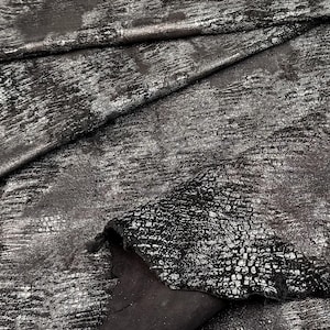 Può includere: Primo piano di un tessuto strutturato nei toni del nero e dell'argento. Il materiale ha un aspetto metallico invecchiato con un motivo screpolato. Il tessuto è piegato, rivelando un lato inferiore opaco più scuro. La trama è irregolare.