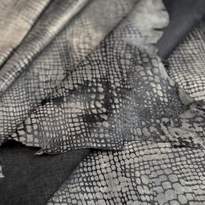 May include: Close-up of a textured, grey and black snakeskin pattern. The leather has a metallic sheen and a scale-like design. The material is folded, revealing the underside of the leather.