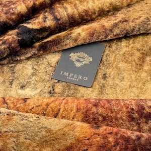 Könnte beinhalten: Nahaufnahme eines Kunstfelltextils mit einem melierten Muster in Braun-, Beige- und Schwarztönen. Der Stoff hat eine weiche, plüschige Textur. Ein Etikett mit dem Text "IMPERO LONDON" ist sichtbar.