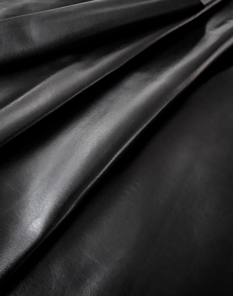 May include: Close-up of black leather with a smooth, textured surface. The leather is folded and draped, creating soft folds and shadows.