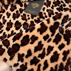May include: Close-up of a plush fabric with a leopard print pattern. The fabric features a tan background with dark brown and black spots. A black tag with gold lettering that reads "IMPERO LONDON" is visible.