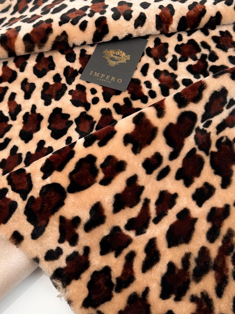 May include: Close-up of a plush fabric with a leopard print pattern. The fabric features a tan background with dark brown and black spots. A black tag with gold lettering that reads "IMPERO LONDON" is visible.