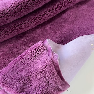 May include: Purple faux fur fabric with a soft, fuzzy texture. The fabric is folded over, revealing a light purple backing.
