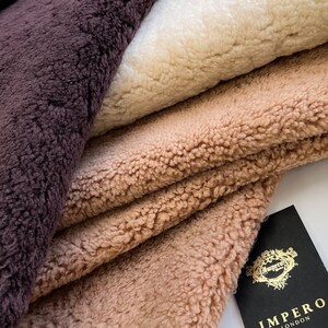 May include: Close-up of four different colored faux fur fabrics: deep purple, cream, light tan, and a medium tan. The fabrics are folded over each other. A black tag with gold lettering that says "IMPERO LONDON" is in the bottom right corner.