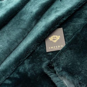 May include: A close-up shot of teal faux fur fabric. The fabric is folded, revealing its texture and depth. A tag with the text "IMPERO LONDON" is attached to the fabric.
