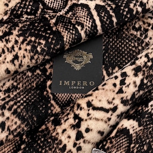 May include: Close-up of a fabric with a snakeskin pattern in shades of black and tan. The fabric is folded, revealing a tag that reads "IMPERO LONDON" in gold lettering on a black background. The fabric has a soft, textured appearance.