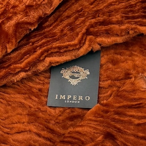 May include: Close-up of a plush, textured, burnt orange fabric. A dark gray tag with gold lettering and a decorative emblem reads "IMPERO LONDON". The fabric has a soft, wavy appearance.
