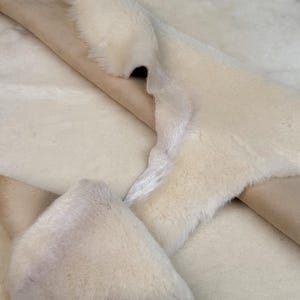 K&ouml;nnte beinhalten: Nahaufnahme eines wei&szlig;en Schaffellteppichs mit weicher, flauschiger Textur. Der Teppich ist in der Mitte gefaltet und zeigt die braune Lederunterseite.