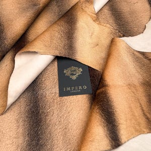 Puede incluir: Primer plano de un trozo de piel de animal marrón y beige. La piel tiene un patrón natural con rayas marrones oscuras y una textura suave. Una etiqueta negra con letras doradas que dice "IMPERO LONDON" es visible.