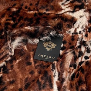 May include: Close-up of a brown and black animal print textile. The textile has a mix of dark brown, black, and white markings. A black tag with gold lettering reads "IMPERO LONDON".