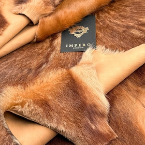 Può includere: Primo piano di una pelle di pelliccia marrone dall'aspetto morbido e strutturato. La pelliccia presenta diverse sfumature di marrone e beige. Un'etichetta nera con scritte dorate recita "IMPERO LONDON". La pelle sembra essere piegata.