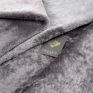 May include: Close-up of a grey faux fur fabric, folded to show its texture. The fabric has a soft, plush appearance. A tag with the text "IMPERO LONDON" is visible.