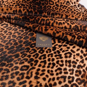 May include: A folded piece of fabric featuring a brown and black leopard print pattern. A small, rectangular, dark grey tag is attached to the material, displaying the gold text IMPERO LONDON with a decorative emblem above the brand name.