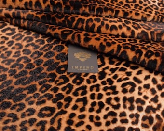 Op de afbeelding: Een gevouwen stuk stof met een bruin en zwart luipaardprintpatroon. Een klein, rechthoekig, donkergrijs label is aan het materiaal bevestigd, met de gouden tekst IMPERO LONDON en een decoratief embleem boven de merknaam.