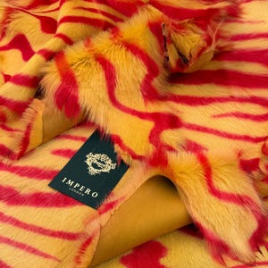 May include: Close-up of a vibrant faux fur fabric with a striking zebra stripe pattern. The fabric features a combination of yellow and red hues, creating a bold and eye-catching design. A tag with the text "IMPERO LONDON" is visible.