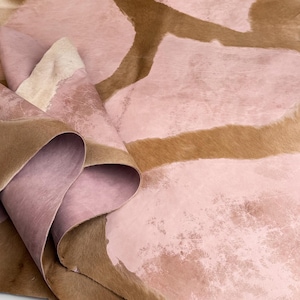 Puede incluir: Una pieza grande de piel animal con una textura de pelaje marrón natural contrastada con grandes parches irregulares de cuero liso y mate de color rosa claro. El material está doblado, revelando el reverso suave y rosado de la piel.