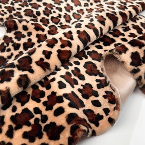 May include: A close-up of a faux fur fabric with a leopard print pattern. The fabric features a beige background with dark brown and black spots. The material is soft and plush, with a visible texture.
