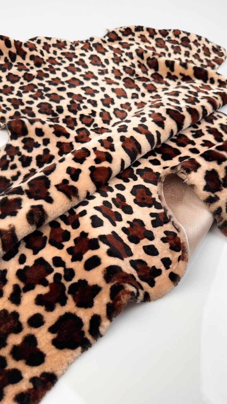May include: A close-up of a faux fur fabric with a leopard print pattern. The fabric features a beige background with dark brown and black spots. The material is soft and plush, with a visible texture.