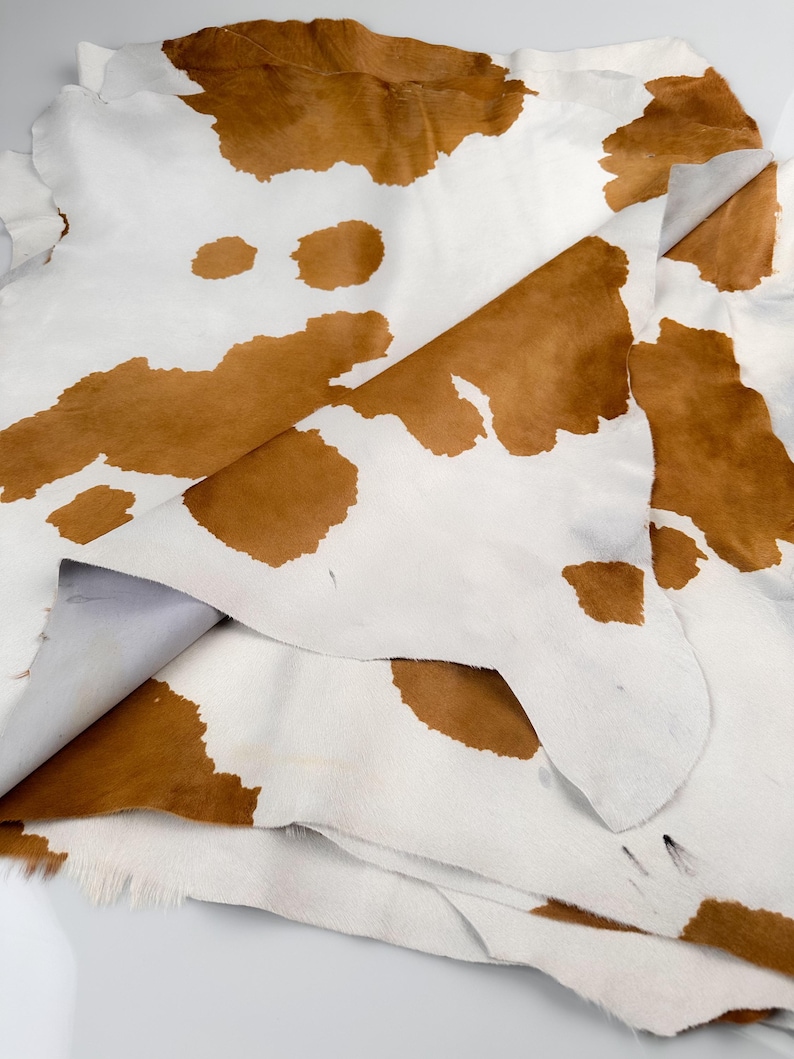 May include: A close-up of several cowhide leather pieces. The hides are white with irregular brown patches. The leather appears soft and pliable, with visible hair and texture. The hides are layered, showing the underside.