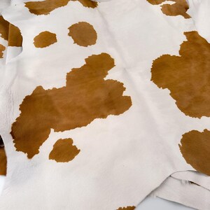 May include: A cowhide rug with a natural pattern of brown spots on a white background. The leather is soft and pliable, with a visible texture. The rug is folded, showing the underside and edges.