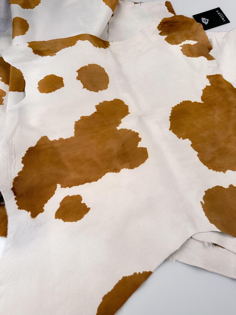 May include: A cowhide rug with a natural pattern of brown spots on a white background. The leather is soft and pliable, with a visible texture. The rug is folded, showing the underside and edges.