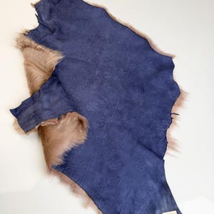 K&ouml;nnte beinhalten: Ein St&uuml;ck indigoblaues Wildleder mit gepr&auml;gtem Blumenmuster, umrandet von hellbraunem Fell. Das Leder hat eine satte Farbe, und das Fell hat ein weiches, nat&uuml;rliches Aussehen. Der Artikel ist wahrscheinlich f&uuml;r Bastel- oder Designzwecke bestimmt.