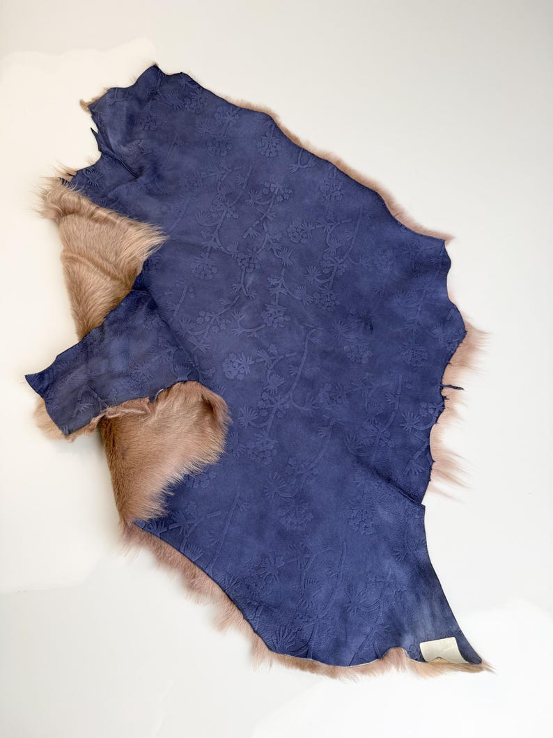 K&ouml;nnte beinhalten: Ein St&uuml;ck indigoblaues Wildleder mit gepr&auml;gtem Blumenmuster, umrandet von hellbraunem Fell. Das Leder hat eine satte Farbe, und das Fell hat ein weiches, nat&uuml;rliches Aussehen. Der Artikel ist wahrscheinlich f&uuml;r Bastel- oder Designzwecke bestimmt.