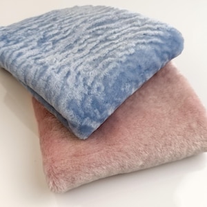 Op de afbeelding: Twee gevouwen vierkanten stof, één lichtblauw met een textuurpatroon, de andere zachtroze. De stoffen hebben een zachte, donzige uitstraling, mogelijk imitatiebont of een vergelijkbaar materiaal. De items zijn netjes opgevouwen.
