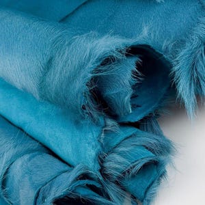 May include: A collection of teal blue cowhide leather pieces with a soft, short-haired fur texture. The material is folded and layered, showcasing the vibrant, saturated colour and the natural, slightly frayed edges of the animal hide material.