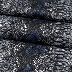May include: A piece of fabric featuring a textured snakeskin pattern in shades of dark blue, black, and metallic silver. The material has a repeating scale-like design with a glossy finish, showing folds and creases across the surface.