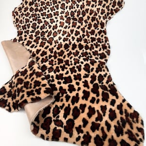 May include: A faux fur pelt with a leopard print pattern. The pelt features a tan base with dark brown spots. The underside of the pelt is a solid tan color. The pelt is irregularly shaped.