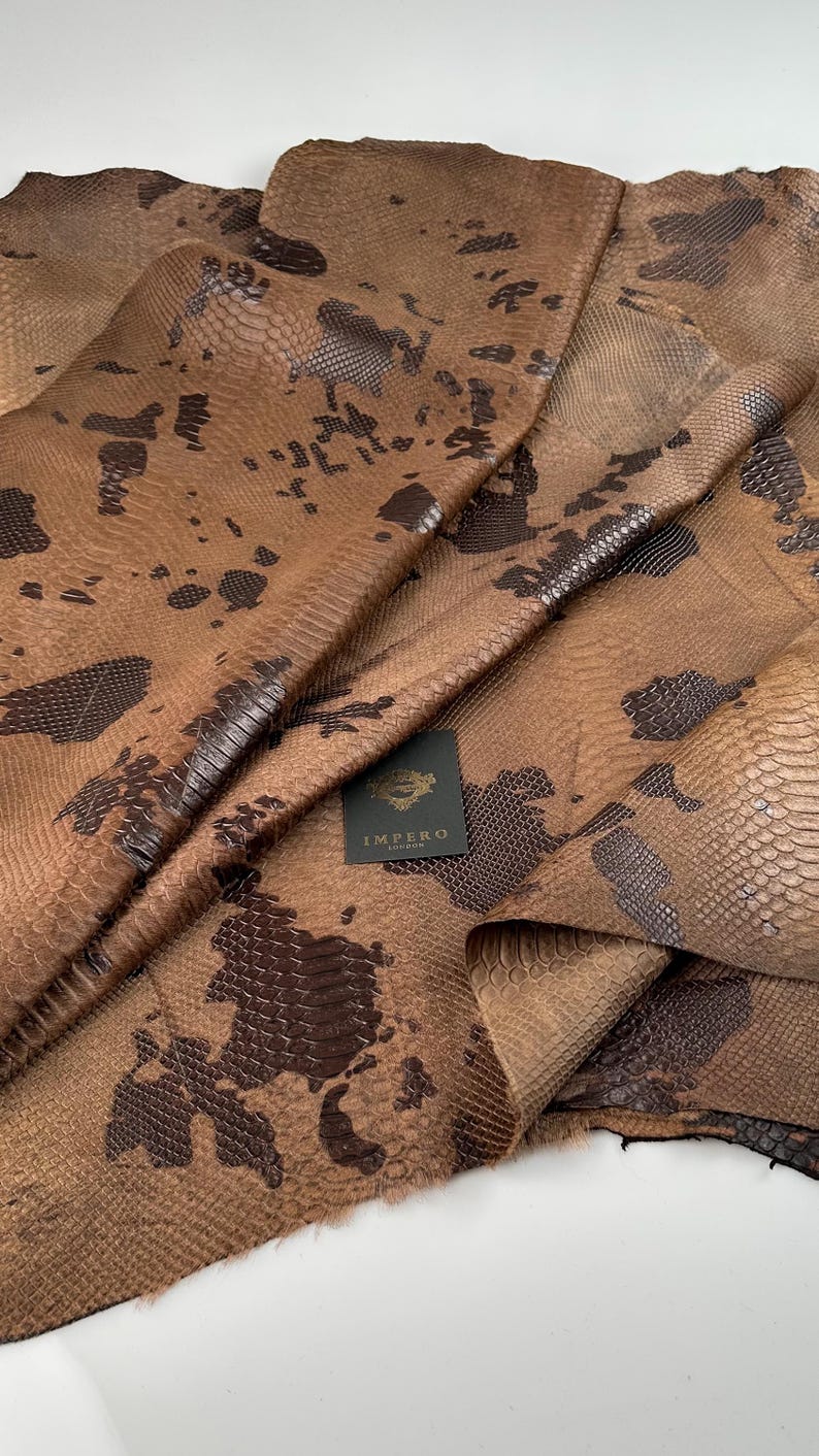 May include: Brown snakeskin leather with a dark brown pattern. The leather is folded and shows the texture of the skin. The leather has a label that says "Impero London".