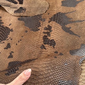 May include: Close-up of a brown snakeskin leather with a dark brown embossed pattern. The leather is folded over, showing the texture and detail.