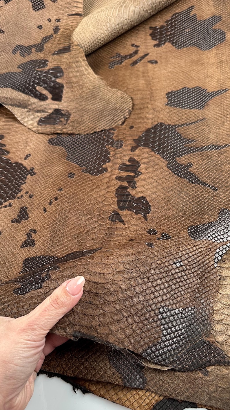 May include: Close-up of a brown snakeskin leather with a dark brown embossed pattern. The leather is folded over, showing the texture and detail.