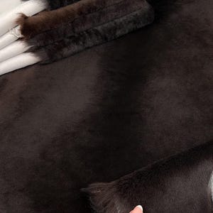 May include: A close-up of a dark brown faux fur fabric. The fabric is soft and fluffy, with a short pile. The fabric is folded in half, and the edges are frayed.