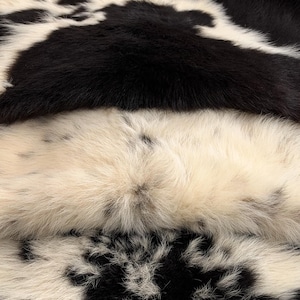 May include: A soft, plush textile featuring a high-pile texture with a distinct black and white cow-print pattern. The material displays dense, layered fur-like fibres in alternating patches of solid black and creamy white tones.