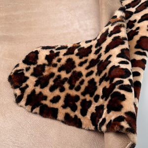 May include: A section of faux fur fabric with a leopard print pattern. The fabric features a tan background with dark brown spots. The fabric is folded over a smooth, light brown material, revealing its underside.