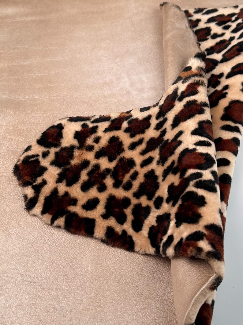 May include: A section of faux fur fabric with a leopard print pattern. The fabric features a tan background with dark brown spots. The fabric is folded over a smooth, light brown material, revealing its underside.