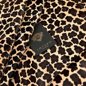 May include: Close-up of a tan and black animal print hide. The hide has a natural pattern of dark brown spots on a lighter tan background. A black tag with gold lettering that reads "IMPERO LONDON" is visible.